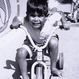 Kids ride trikes by an Aboriginal mural, no date